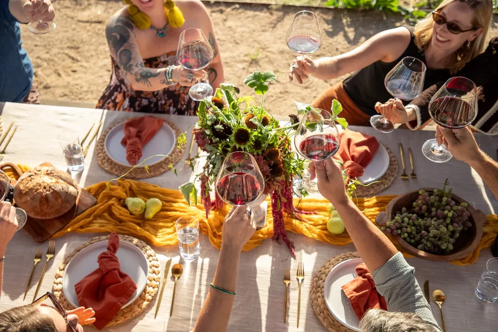 Guests raising glasses of red wine over a long harvest table with bread, grapes and pears.