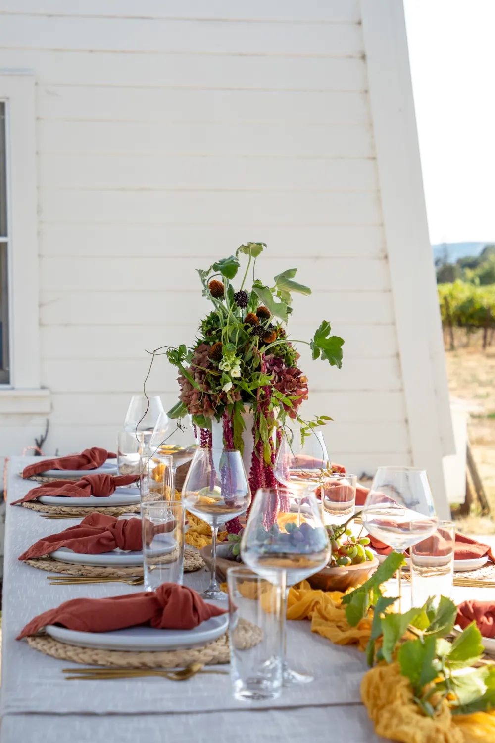 A table set against a white barn wall with a tall floral centerpiece.