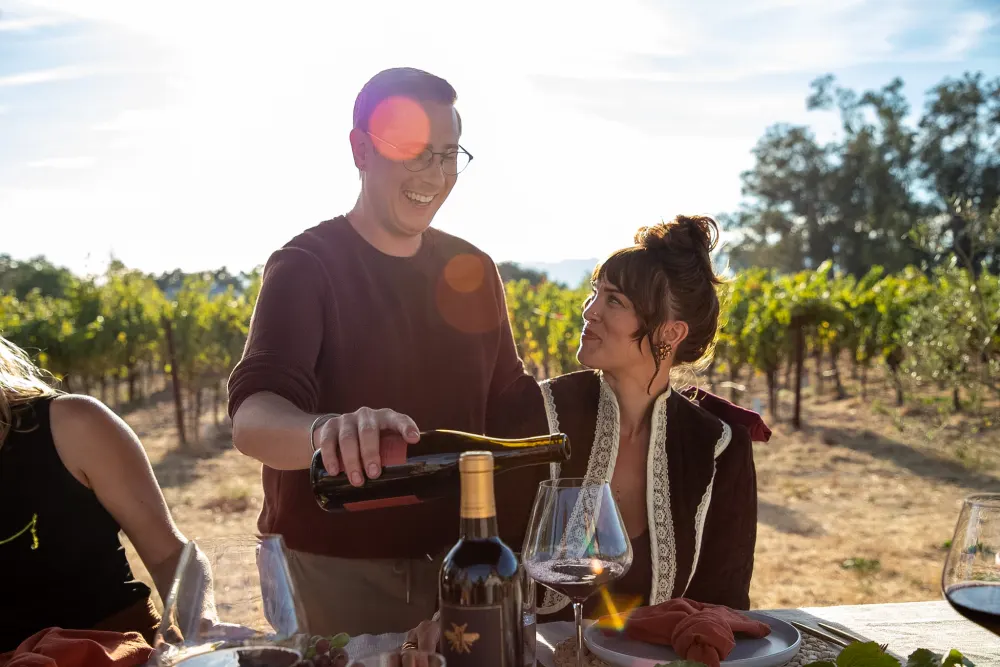 A guest pouring red wine for a friend at a sunlit vineyard table.