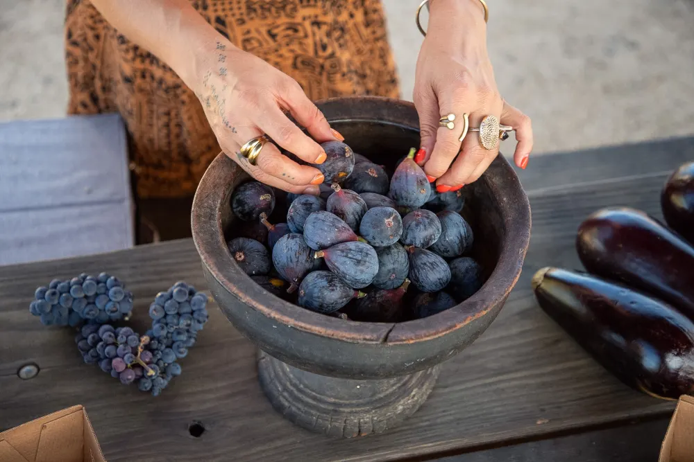 Hands arranging fresh figs in a wooden pedestal bowl.