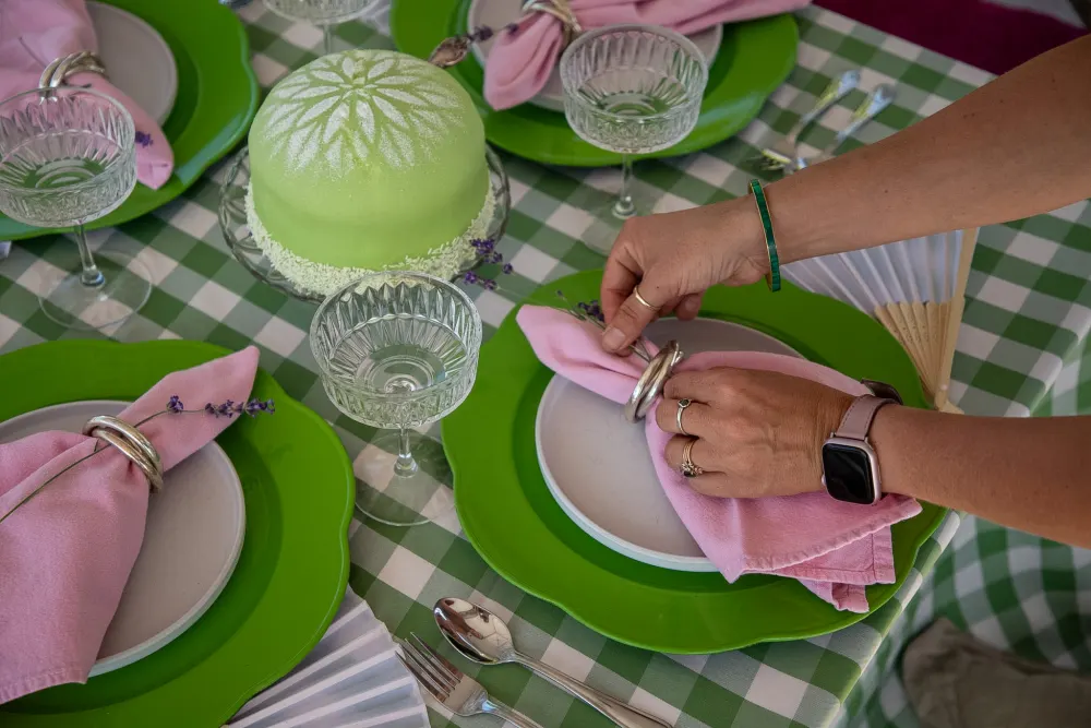 A hand placing a pink napkin on a green gingham picnic setting.