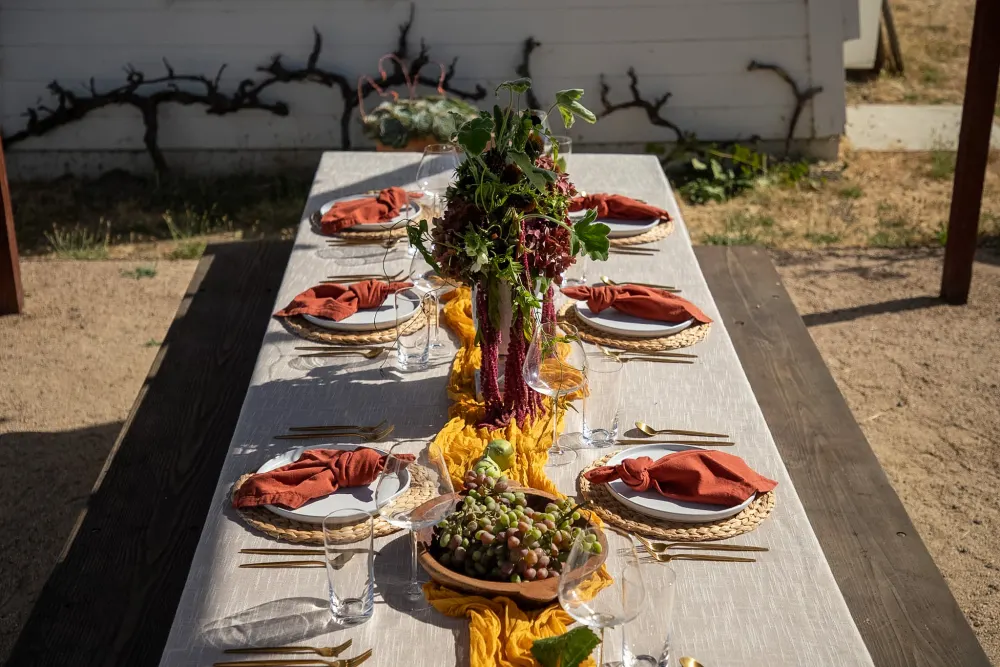 A long vineyard table fully styled with centerpieces, woven chargers and rust napkins.
