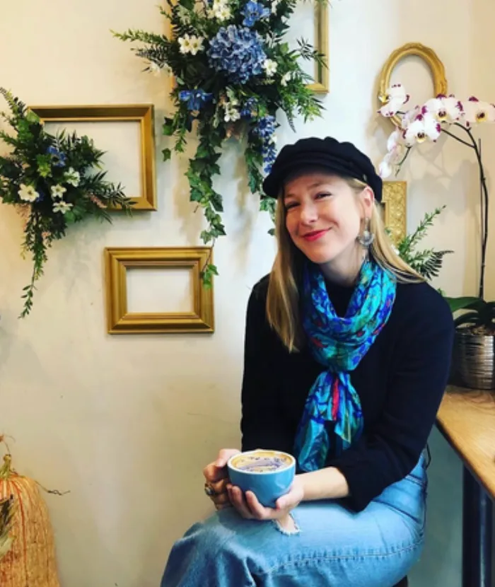 Kelsey Knego smiling with a coffee cup among gilded frames and florals.